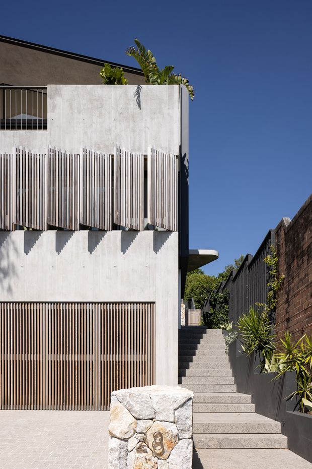 Exterior view of a contemporary multi-level urban residence with a pale rendered or concrete facade, extensive vertical timber screening, a dark roof edge, and a stairway rising beside a landscaped boundary wall. The facade combines solid geometric volumes with sun-shading slats, while tropical planting and a clear blue sky create a bright outdoor setting.