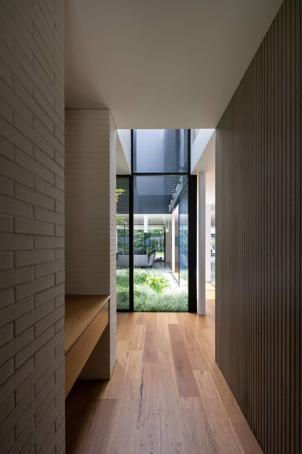 A contemporary residential hallway or entry passage with warm timber flooring, a white painted brick wall and integrated timber bench on the left, and a dark vertical timber slat wall on the right. Full-height black-framed glazing at the end opens views toward a landscaped internal courtyard and brings in natural light. The corridor has a clean, linear composition and a restrained neutral palette.