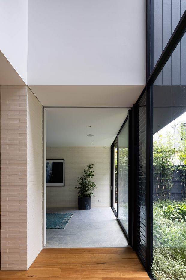 A minimalist residential hallway or transitional gallery space with pale horizontal brick walls, a smooth white ceiling, polished grey concrete flooring, and warm timber flooring in the foreground. Full-height black-framed glazing runs along the right side, overlooking a planted garden. A large potted plant, framed artwork, and a small rug provide restrained interior detailing. The space is bright, calm, and strongly connected to the garden through the glazing.