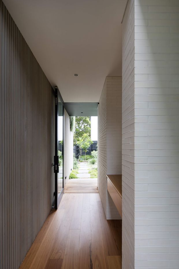 A narrow, covered residential passage leading from an interior entry toward a landscaped garden. The corridor has warm timber flooring, a dark vertical timber slat wall on the left, white painted brick walls on the right, recessed ceiling lighting, full-height glazing, and a built-in timber bench or joinery recess. The space is linear, calm, and naturally lit, with the garden visible at the far end.
