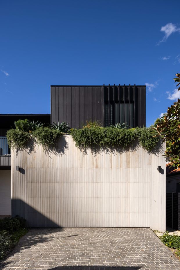 Exterior view of a contemporary multi-level residence, featuring a tall light-toned vertically ribbed facade, dark upper volume with vertical cladding and narrow windows, a planted green screening layer, and a grey brick-paved driveway under a clear blue sky.