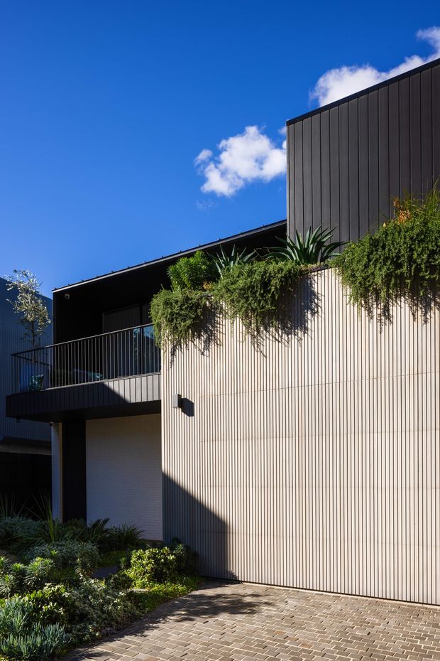 Exterior architectural photograph of a contemporary multi-level residence featuring dark vertical cladding, a pale vertically ribbed wall, balcony with slim metal balustrade, garage door, landscaped planting, and a paved driveway. Strong sunlight creates crisp shadows against a vivid blue sky.