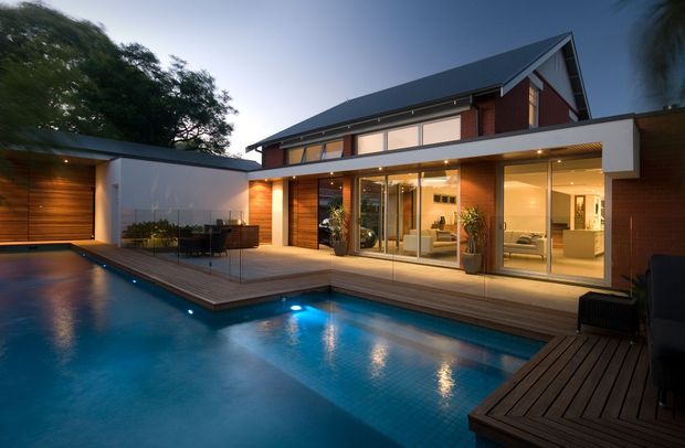 Twilight exterior view of a contemporary multi-level residence centred around a swimming pool and timber decking. The home features a steep gabled roof, dark roofing, red-brown brick and warm timber-clad walls, extensive floor-to-ceiling glazing, and a covered outdoor entertaining area. Interior living spaces are visible through the glass, with integrated lighting creating a warm glow.