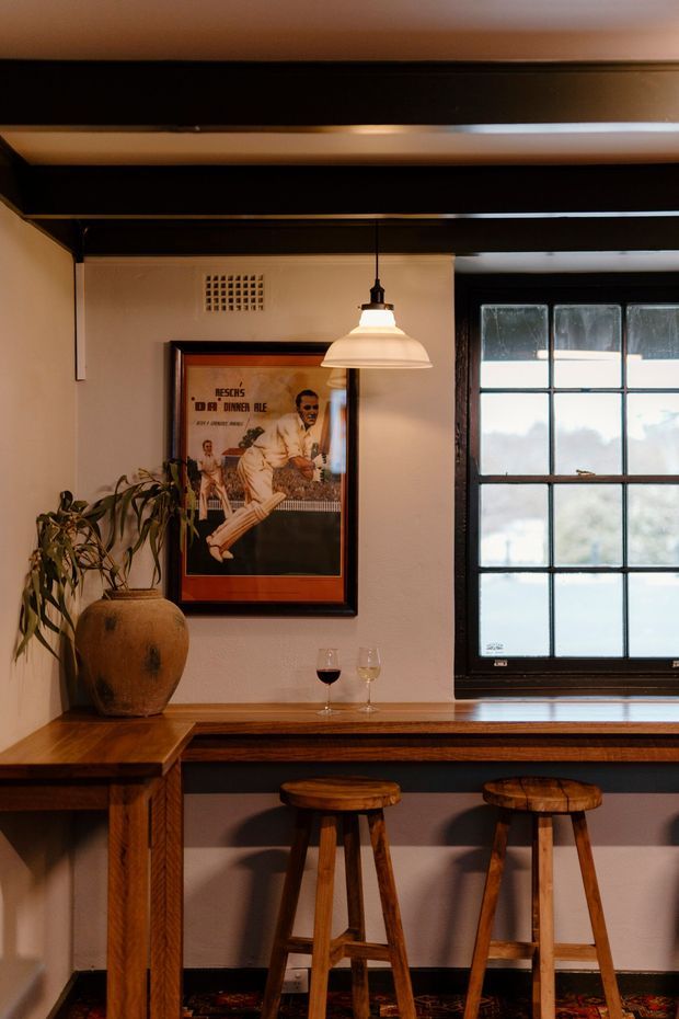 A warm, moody interior bar nook with a timber counter and two wooden stools beneath a pendant light. Dark exposed ceiling beams, pale plaster walls, a black-framed multi-pane window, framed artwork, a ceramic vase with foliage, and wine glasses create a vintage, intimate atmosphere.