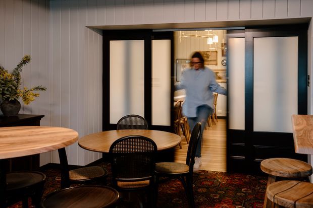 A warm, eclectic dining area viewed through wide black-framed glazed doors. The room features a round timber dining table with black cane-back chairs, additional timber stools and tables, vertical wall panelling, a patterned dark rug, and a softly illuminated adjoining space beyond the open doors.