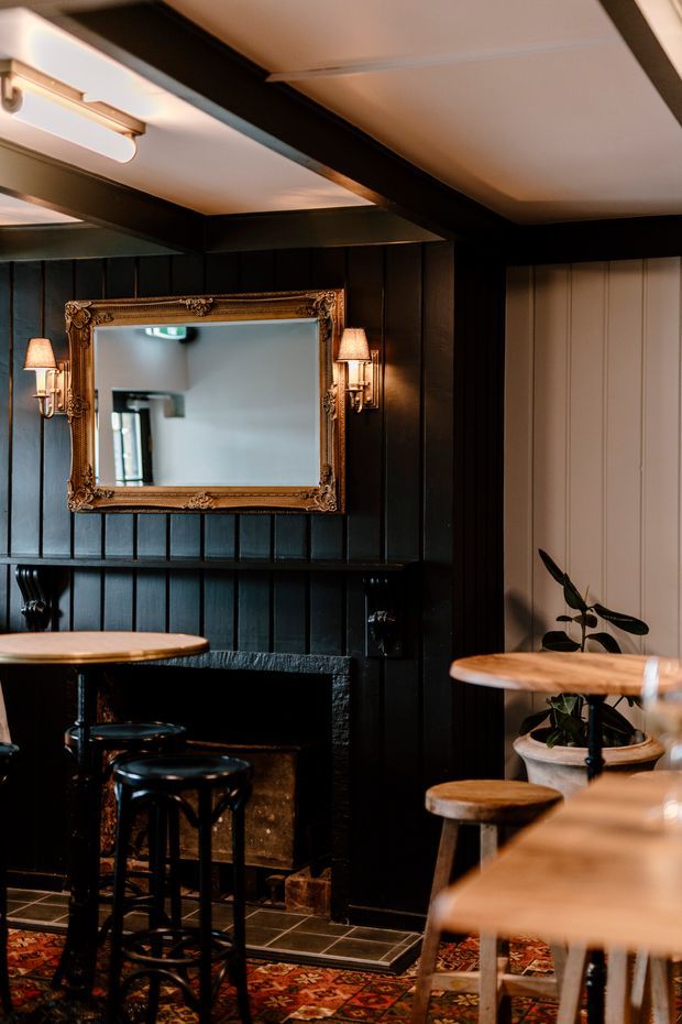 A moody, intimate bar or dining nook with black vertical timber wall panelling, a dark tiled fireplace surround, and an ornate gold-framed mirror above the hearth. Small timber-topped tables and stools occupy the room, with wall sconces providing warm lighting. A patterned rug and potted plants add texture, while a pale panelled door or wall section contrasts with the dark interior.
