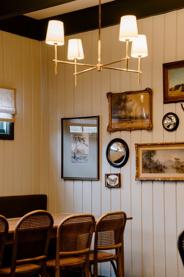 A warm, intimate dining room with vertical cream-painted wall panelling, a dark ceiling, and a brass multi-arm pendant chandelier with fabric shades. A timber dining table is surrounded by cane-back chairs, while the walls display an eclectic arrangement of framed landscape artwork, round mirrors, and a small framed butterfly.