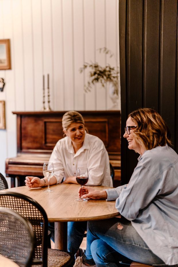 Warm, candid interior scene of two people seated at a round wooden table enjoying drinks. The space features a dark timber upright piano, white vertical wall panelling, black-framed detailing, woven cane dining chairs, and soft natural styling with greenery.