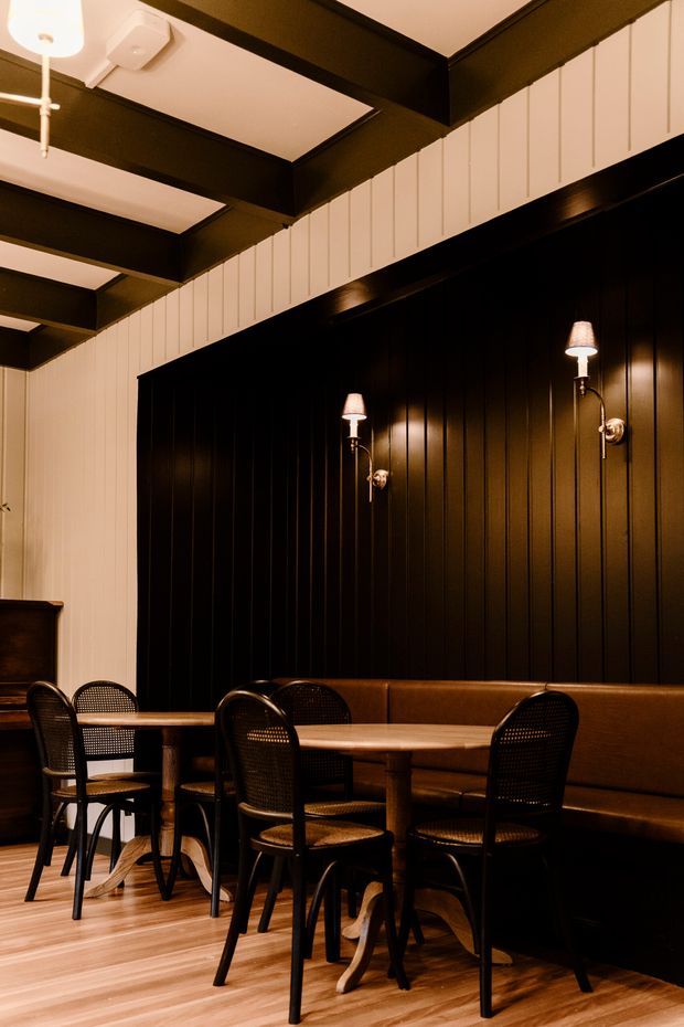 A warmly lit commercial dining interior with dark vertical timber wall panelling, built-in brown banquette seating, round wooden tables, and black bentwood-style cane chairs. Brass wall sconces and black exposed ceiling beams contrast with light vertical wall cladding and timber flooring, creating an intimate restaurant or café atmosphere.