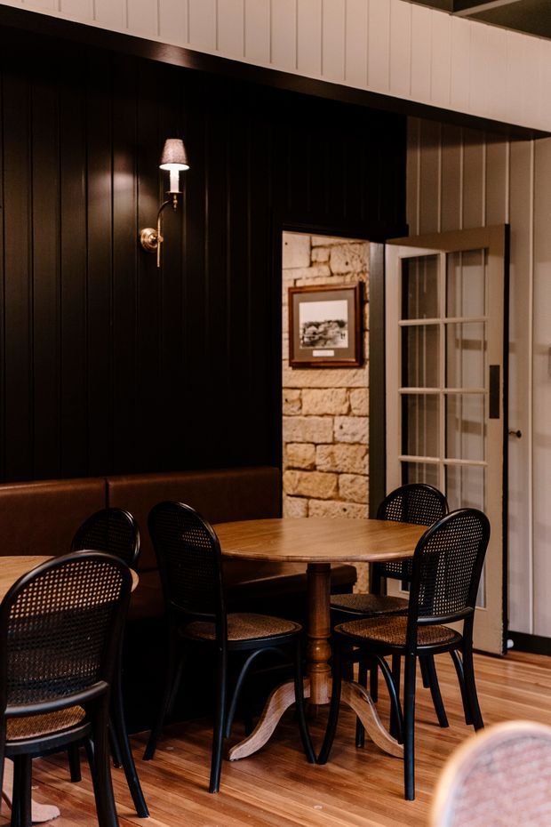 A warmly lit dining area with a round timber pedestal table and black woven-back chairs, set against dark vertical wall panelling and a leather banquette. A white-framed glazed French door, exposed light stonework, framed artwork, and timber flooring add character and contrast.