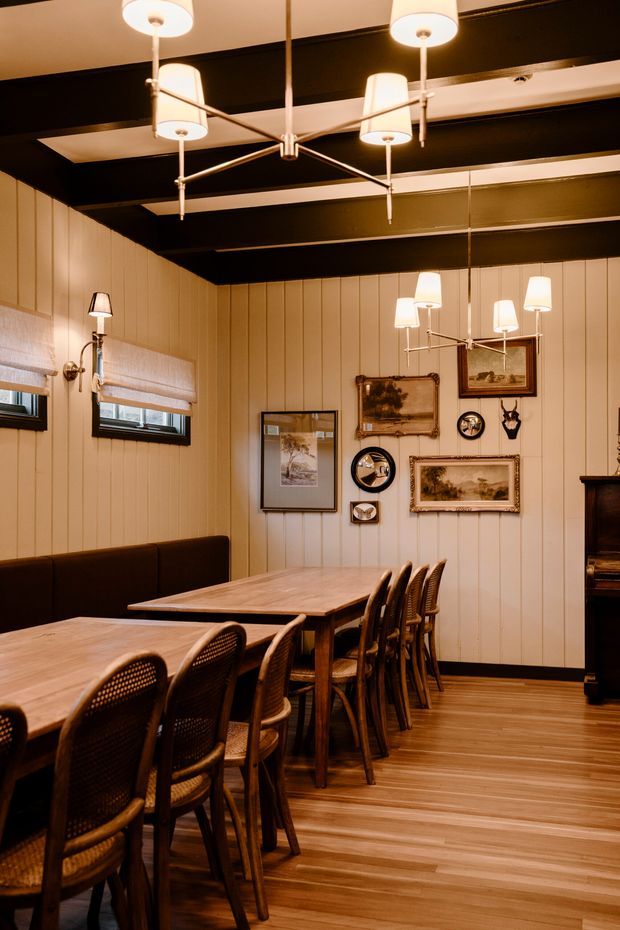 A warm, refined dining room with long timber dining tables and rows of woven-back timber chairs. Vertical cream wall panelling, timber-look flooring, framed artwork, and multiple contemporary chandelier-style pendant fixtures create a cozy restaurant-like atmosphere. Dark exposed ceiling beams provide strong contrast against the pale walls.
