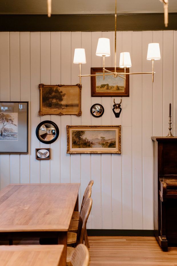 A warm, traditionally styled dining room with a large light-wood dining table and wooden chairs. The walls are clad in vertical painted panels and decorated with framed landscape artwork, a round mirror, a small butterfly picture, and a mounted antler motif. A brass-toned linear chandelier with white shades hangs above the table, while a dark upright piano and decorative candlestick occupy the right side of the room. Timber flooring and dark trim complete the refined, heritage-inspired interior.