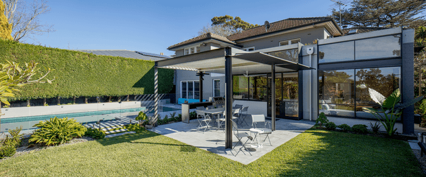 Exterior view of a landscaped residential courtyard and outdoor entertaining area. A paved patio with a black-framed pergola and outdoor dining furniture sits beside a swimming pool, with expansive glazed doors connecting to the house. The home has a light rendered facade, dark-framed glazing, and a tiled hipped roof, surrounded by manicured lawn and planting in bright daylight.