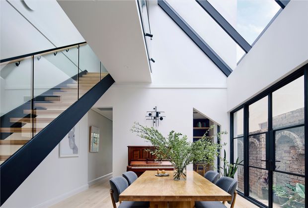 Bright contemporary dining area within a double-height interior, featuring a large timber dining table, upholstered dining chairs, a sculptural wall clock, and a timber stair with dark painted stringer and clear glass balustrade. Large black-framed glazing and overhead skylights bring in abundant daylight, while exposed brickwork is visible outside.
