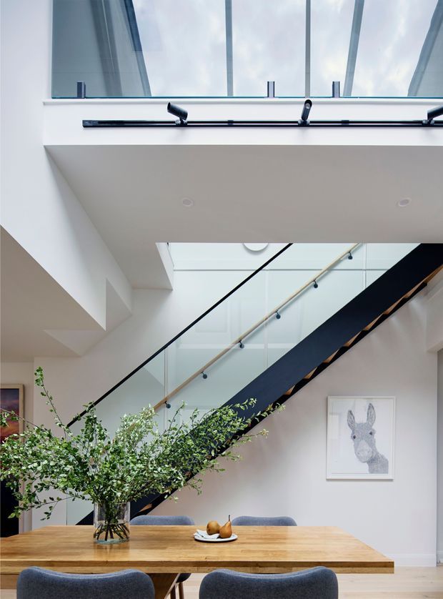 Bright contemporary dining area beneath a prominent open stairway. A large timber dining table with upholstered chairs sits in the foreground, accompanied by a tall leafy branch arrangement and simple fruit bowl. The stair features a dark structural stringer, timber handrail, and clear glass balustrade, while a large skylight or roof glazing brings daylight into the double-height space. White walls and ceiling create an airy, refined interior, with a framed animal artwork on the wall.