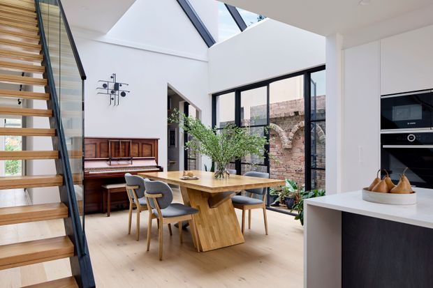 Bright contemporary open-plan dining area beside a modern kitchen, with a timber dining table and chairs, upright piano, floating timber staircase, skylights, and expansive black-framed glazing overlooking an exposed brick courtyard wall. The space combines white walls, pale timber flooring, dark cabinetry, and abundant natural light.