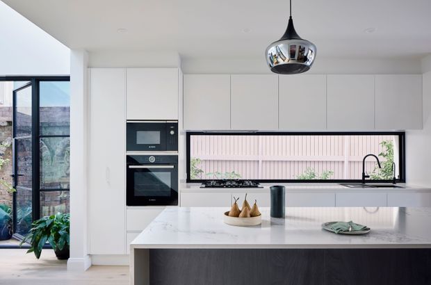 Bright contemporary kitchen with extensive white flat-panel cabinetry, integrated black appliances, a large pale stone island, and a long horizontal window above the sink. A dark timber-toned island base contrasts with the light surfaces, while a polished chrome pendant adds a focal point. Glazing at left opens toward a planted outdoor area.