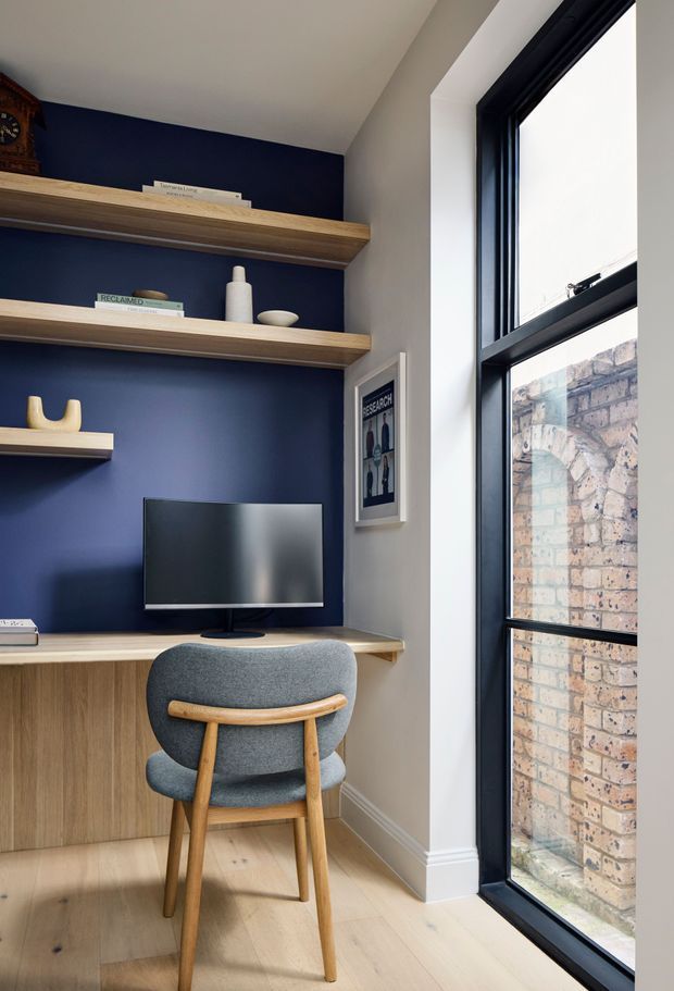 Compact home office nook with a built-in timber desk and open shelving set against a deep navy feature wall. A monitor, framed artwork, books and simple decorative objects occupy the joinery, while a grey upholstered timber-framed chair faces the desk. Full-height black-framed glazing on the right overlooks a brick wall, bringing in natural light. The space has pale timber flooring, white trim and a clean, tailored contemporary feel.