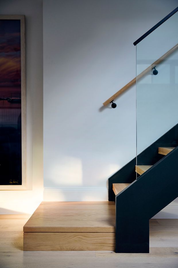 Interior view of a contemporary stairway with pale timber flooring and stair treads, a strong black stringer and balustrade, clear glass guarding, and a simple timber handrail mounted to a white wall. A framed artwork is visible at left, with natural light creating soft shadows across the stair hall.