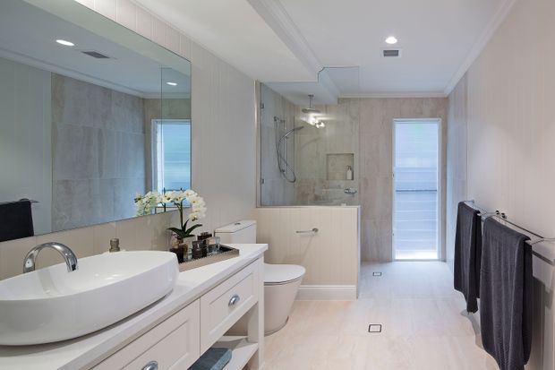 Bright contemporary bathroom with a long white vanity, vessel basin, large wall mirror, toilet, and a glass-enclosed walk-in shower with pale stone-look wall and floor finishes. White cabinetry, chrome fittings, recessed lighting, and a freestanding bath create a clean, spacious appearance.
