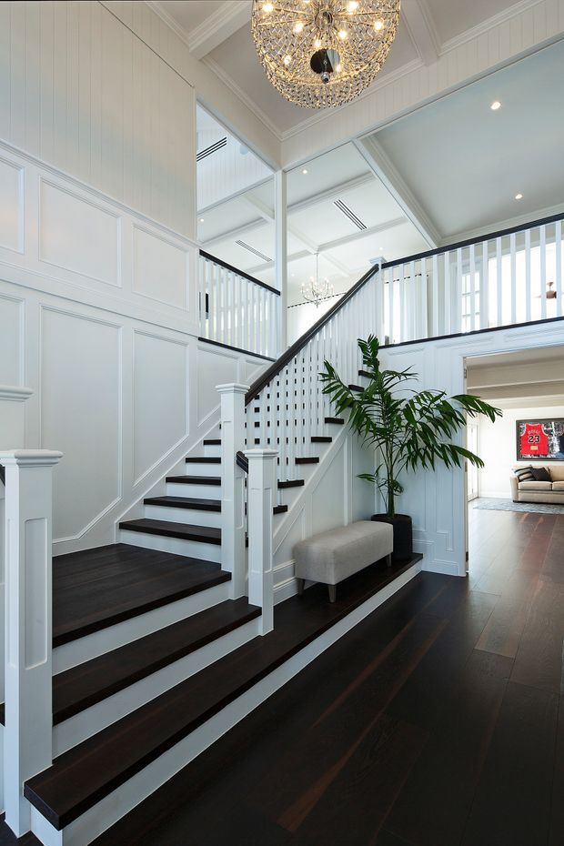 A bright, formal residential stairway and landing with dark timber stair treads and flooring, white painted panelled walls, white vertical balustrades, and a black handrail. A large decorative crystal pendant hangs above the stairwell, while a potted palm and upholstered bench sit on the landing. An open doorway reveals part of an adjoining living room in the background.
