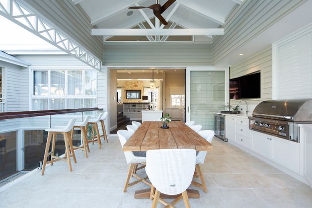 Bright, spacious enclosed outdoor entertaining room with a large timber dining table, white upholstered chairs, a bar-height counter and stools, built-in white cabinetry, a built-in barbecue, and a wall-mounted television. The room has extensive glazing, pale horizontal wall cladding, exposed white roof trusses, a pitched ceiling, and a ceiling fan, with a kitchen visible beyond the open doors.