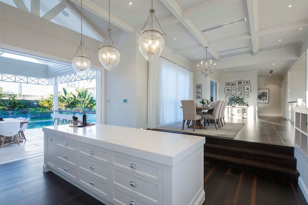 Bright, spacious open-plan kitchen and dining area with a large white island featuring extensive drawer cabinetry, pendant glass lighting, dark timber flooring, and a raised dining platform. Large glazed openings and skylights bring in abundant daylight, with a pool and landscaped outdoor area visible beyond.