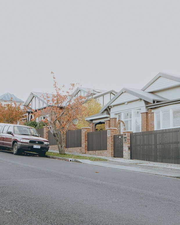 Street-level view of a quiet suburban residential neighbourhood with closely spaced detached homes. The foreground house has a light weatherboard facade, gabled roof, brick piers and boundary walls, white plantation shutters, and a dark vertical-slat timber fence and gate. Additional gabled homes, autumn deciduous trees, paved footpaths and a parked vintage car are visible along the street.