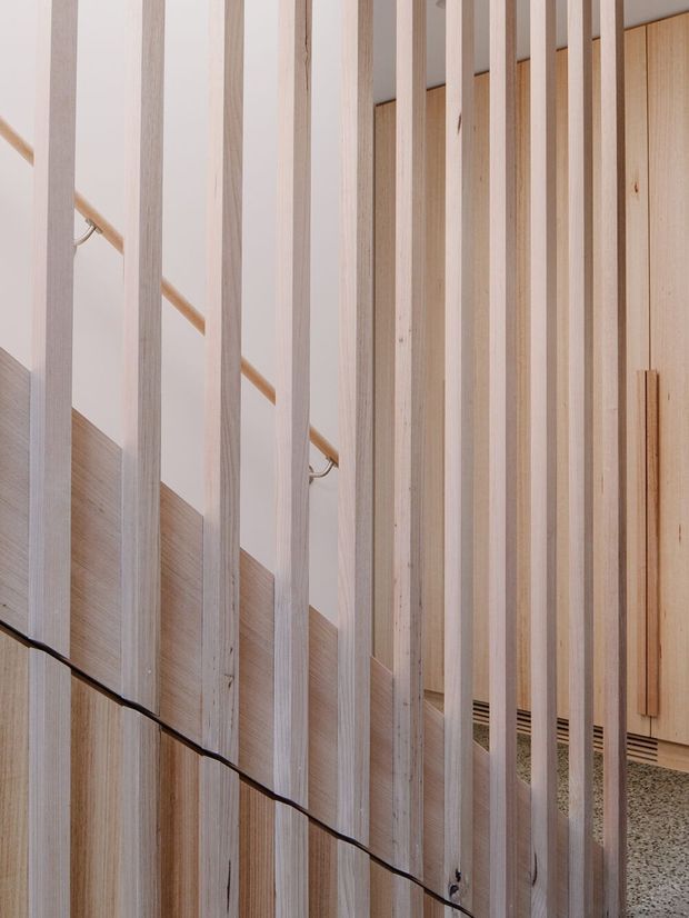 Close-up interior view of a timber slatted screen or balustrade, with evenly spaced pale vertical wood battens partially concealing a staircase and handrail. Light timber cabinetry or wall panelling is visible behind, along with a terrazzo-like floor. The composition highlights warm natural wood grain, linear detailing, and filtered visibility through the screen.
