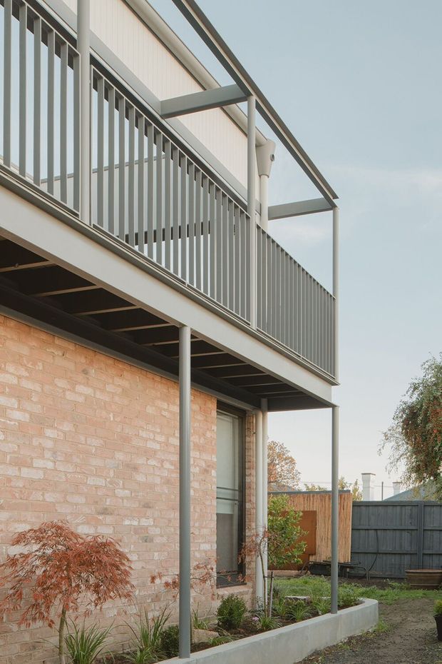 Exterior architectural photograph of a contemporary two-level brick building with a raised balcony or terrace. The balcony has slender grey metal vertical balustrades, exposed steel framing, and a simple flat canopy or roof edge supported by slim posts. Pale brick walls, dark structural elements, a timber privacy fence, and a planted garden bed are visible at ground level.