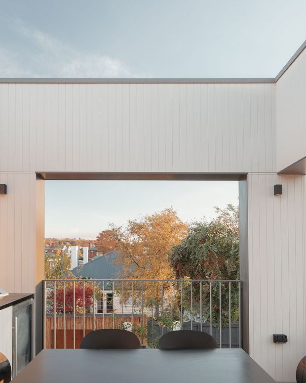 A contemporary covered outdoor dining area framed by pale vertical cladding and a large rectangular opening. A dark dining table and chairs sit beside a metal balustrade, overlooking trees and neighbouring homes in an autumnal suburban setting. The space has clean geometric lines, integrated wall lighting and a sheltered, open-air character.