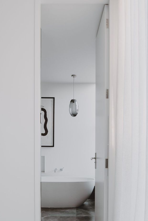 A minimalist white bathroom viewed through an open doorway, featuring a freestanding oval bathtub, grey stone-look floor tiles, a clear glass pendant light, white walls, a wall-mounted tap, and a framed abstract artwork. White door trim and a sheer curtain frame the view.