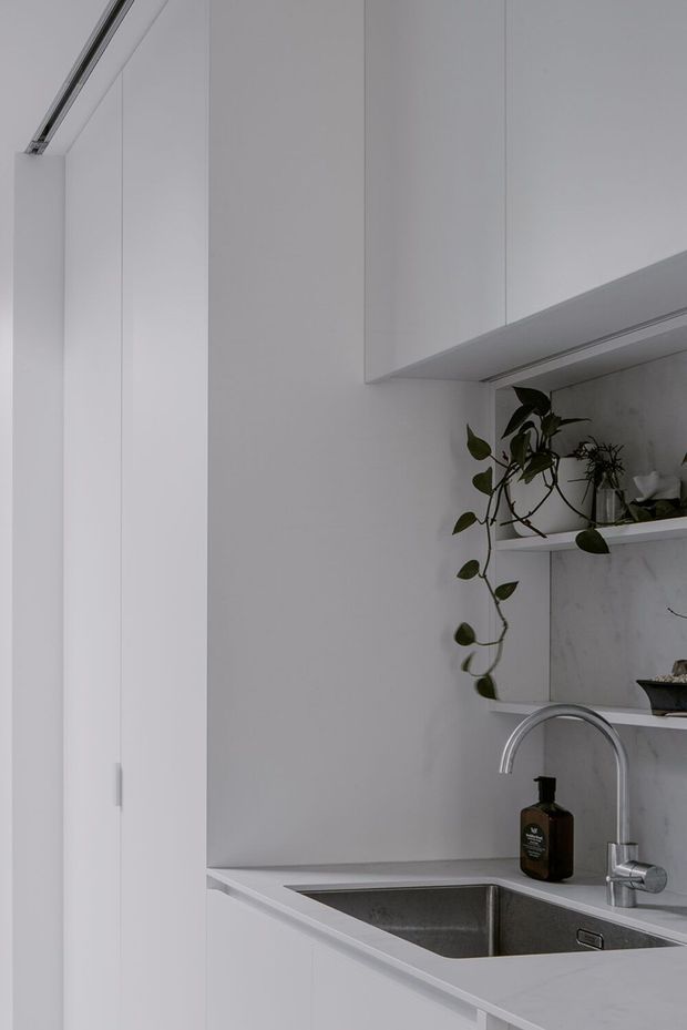 A close-up of a compact, minimalist white kitchen featuring handleless full-height cabinetry, a stainless-steel undermount sink with curved chrome tapware, a pale veined stone backsplash and benchtop, and open shelving displaying potted plants. The space is softly lit with a restrained monochromatic palette and clean built-in detailing.