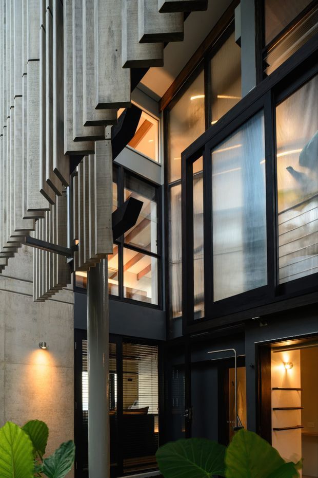 A dramatic multi-level interior courtyard or atrium with exposed concrete walls and a sculptural cantilevered concrete staircase. Black-framed glazing with translucent ribbed panels encloses adjoining spaces, while warm interior lighting, timber accents, and indoor tropical planting soften the industrial architectural composition.