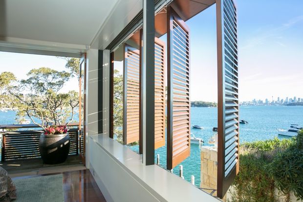 A bright coastal balcony or terrace overlooking a turquoise harbour, with timber decking, dark-framed glazing, horizontal louvred screens, a black planter, and a covered outdoor seating area. The city skyline and boats are visible across the water.