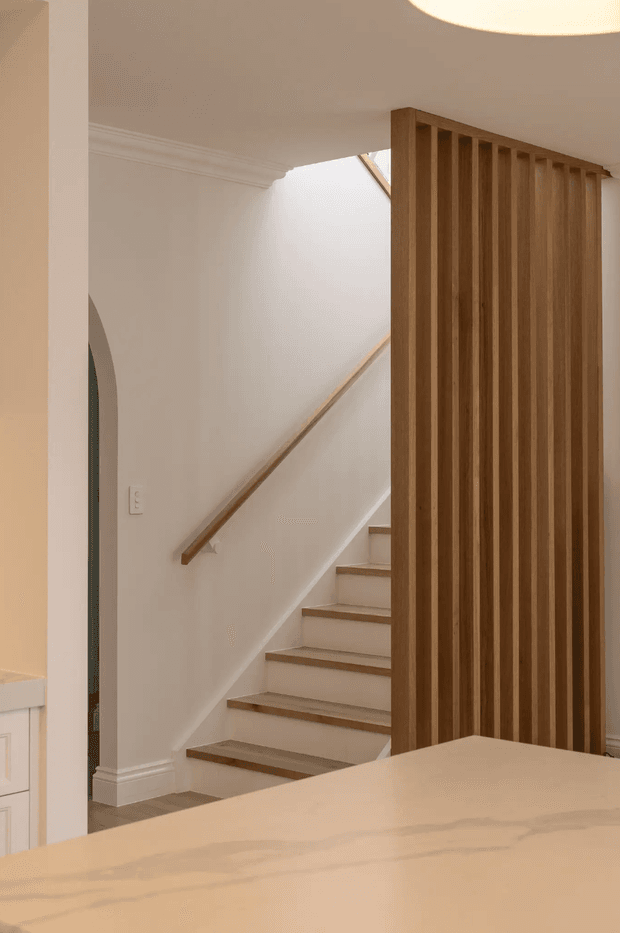 Warm, refined interior view of a residential stairway beside a tall vertical timber slatted screen. White walls with traditional cornice and skirting frame timber stair treads and a simple wall-mounted handrail. Part of a pale stone or timber kitchen island is visible in the foreground, with soft recessed or pendant lighting overhead.