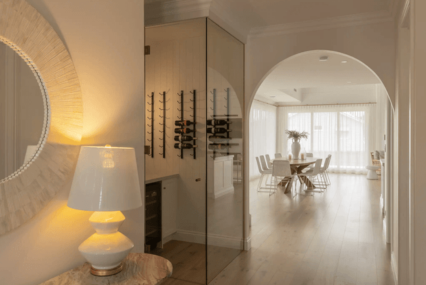 Warm, refined interior view looking from an entry or hallway toward a bright dining area. The foreground features a large textured round mirror, illuminated table lamp, marble-topped console, and a built-in wine storage wall with dark timber flooring. An arched opening leads to a light-filled dining room with a round table, upholstered chairs, sheer curtains, and a large window. White panelled walls, detailed ceiling cornices, and traditional joinery create a polished classic character.