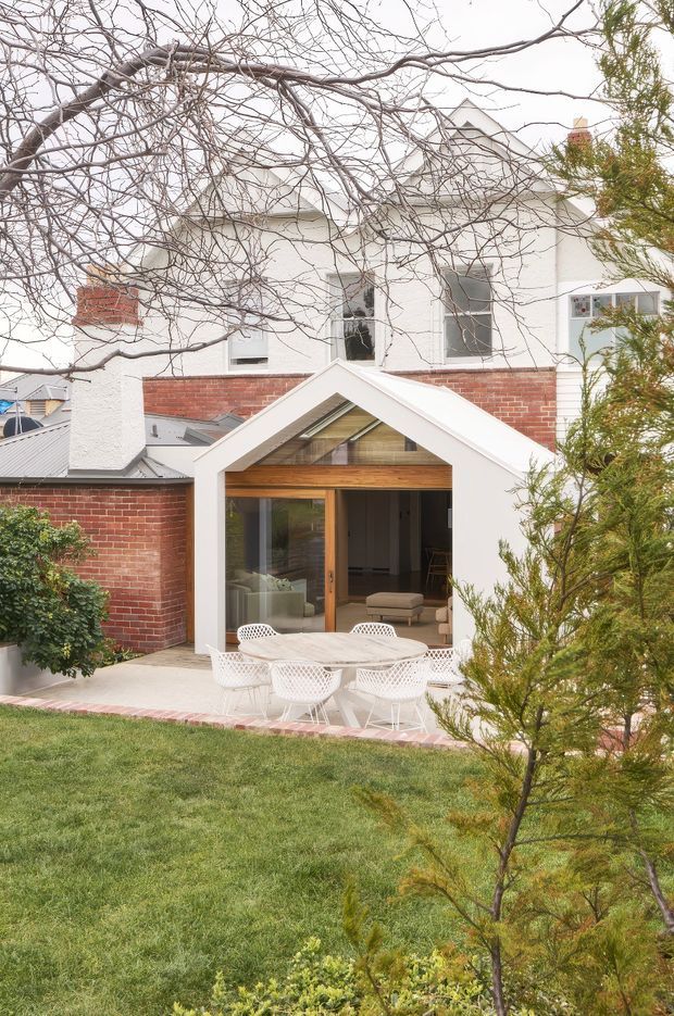 Exterior view of a renovated two-storey brick-and-white-rendered house with a prominent gabled rear extension. The extension has a white gable roof and trim, exposed warm timber framing beneath the gable, and large timber-framed glazed sliding doors opening onto a paved outdoor dining terrace. White outdoor dining chairs and a round table sit beside the lawn, with established garden planting and mature trees in the foreground.
