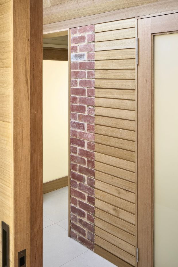Close-up interior view of a doorway and feature wall. The composition combines warm timber door frames and ceiling lining with a red exposed-brick vertical strip and horizontal timber slatted wall cladding. A timber-framed frosted-glass door is partially open, revealing a simple tiled-floor room beyond.