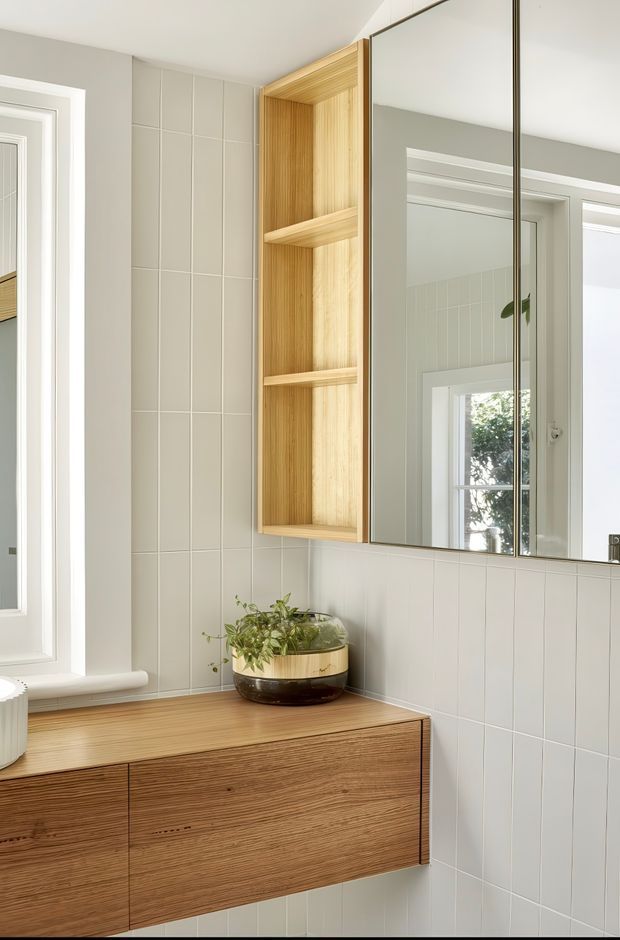 A bright, compact bathroom vanity area with pale vertically gridded wall tiles, a wall-mounted timber vanity with integrated drawers, an open timber shelving niche, and mirrored cabinet doors. A small planter sits on the vanity, while a window and reflected doorway bring in natural light. The design is clean, warm, and contemporary with a strong wood-and-white palette.