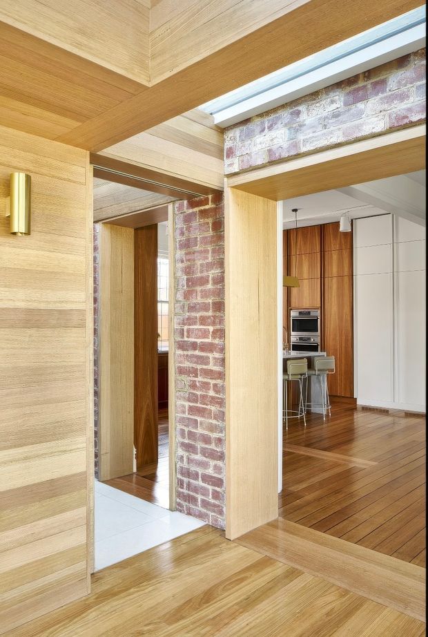 Interior view of a warm, timber-lined home with a central exposed red-brick structural wall and timber-framed openings. The image looks through a circulation area toward a compact kitchen with integrated appliances, wood cabinetry, a small island or breakfast bar, and stools. Timber flooring, ceiling beams, wall panelling, and recessed doorways create a cohesive refined modern interior, with a light tiled floor visible in an adjacent space.