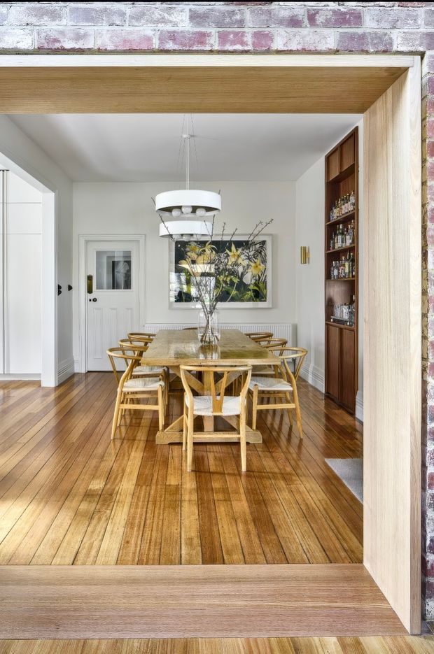 Bright dining room viewed through a wide timber-lined opening, featuring a long rectangular dining table with woven cane dining chairs, polished timber floorboards, white walls with vertical panelling, a built-in dark timber drinks cabinet, framed artwork, and a contemporary pendant light. Exposed painted brick is visible around the opening, creating a blend of traditional and updated detailing.