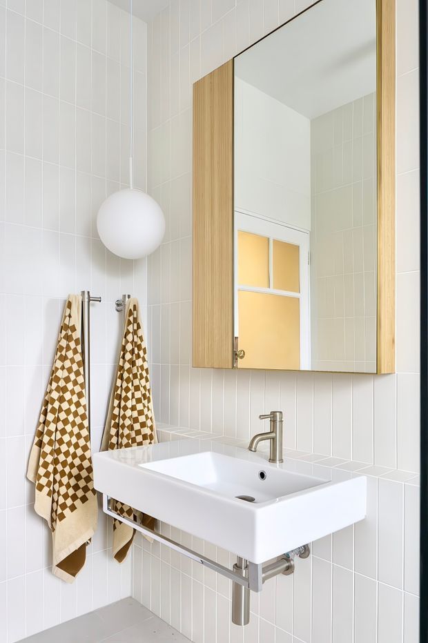 A bright, compact bathroom featuring pale vertically aligned wall tiles, a wall-mounted white rectangular basin with exposed chrome plumbing, a chrome single-lever tap, and a large mirrored cabinet with light timber side panels. A spherical white pendant light and patterned towels add decorative detail.