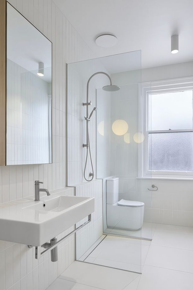Bright, compact contemporary bathroom featuring a wall-mounted white basin, frameless glass walk-in shower with brushed metal fixtures, wall-hung toilet, large mirror, white vertical wall tiles, pale floor tiles, and frosted window glazing. The design is clean and minimalist with strong white tones and abundant natural light.