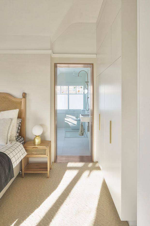 Bright, calm bedroom viewed toward an adjoining bathroom. The bedroom features a cane/rattan bedhead, patterned bedding, a small timber bedside table, warm neutral carpet, white walls with decorative cornices, and full-height white built-in wardrobes with slim brass handles. The bathroom beyond has pale blue-green walls, light tiled surfaces, a shower, freestanding or built-in bath, and strong natural light through a window.