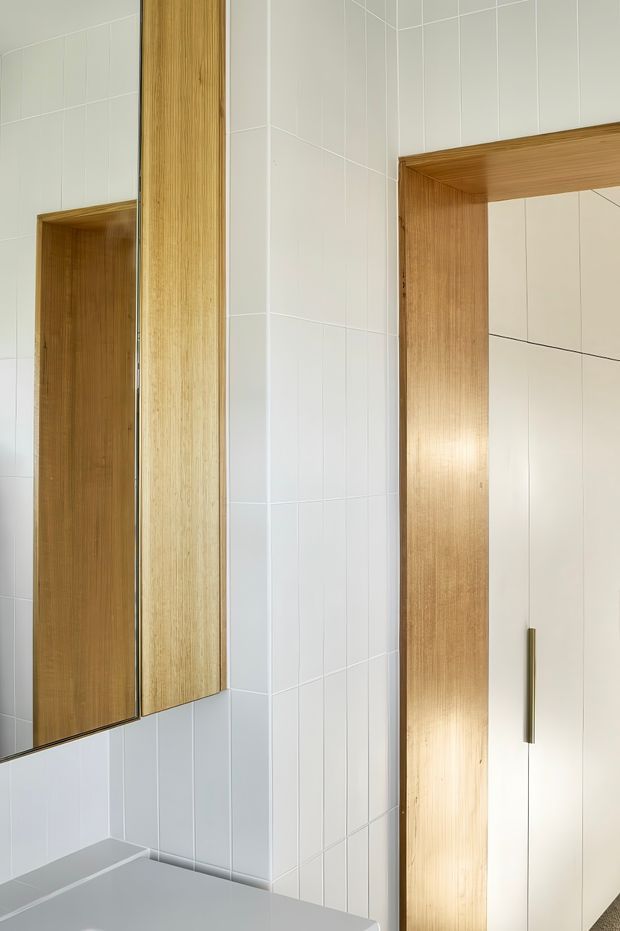 Close-up view of a contemporary bathroom vanity area with white tiled walls, a large mirrored cabinet with warm timber framing, and a matching timber-framed recessed storage or doorway. The space has clean lines, integrated joinery, and a restrained light neutral palette.