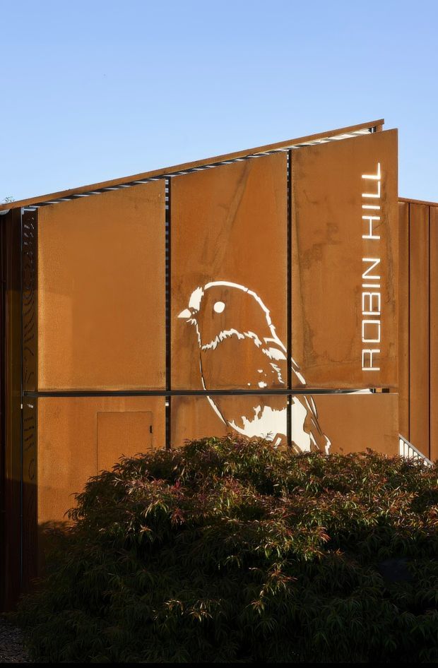 Exterior view of a contemporary building clad in weathered rust-toned steel panels, with black structural framing, a vertical illuminated 'ROBIN HILL' sign, and a large white bird mural cut into or applied to the facade. Dense landscaping obscures the lower portion, while a clear blue sky forms the background.