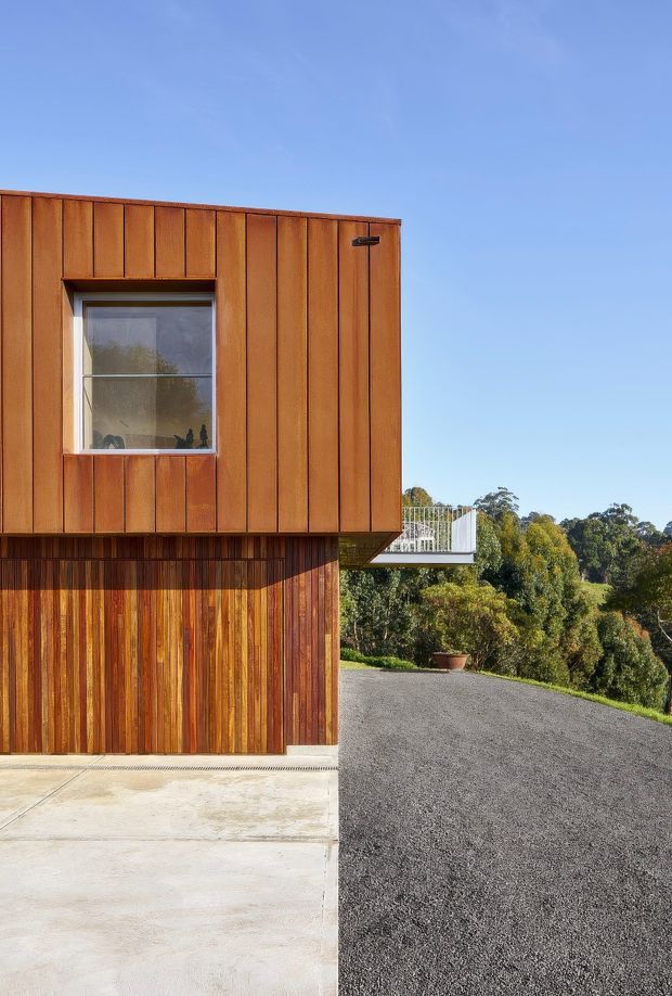 Exterior architectural photograph of a compact contemporary building clad in vertical warm-toned timber boards. The view shows a projecting upper volume with a recessed window, a timber-clad lower level, a flat roofline, concrete paving, and a dark gravel driveway. A small elevated deck with a pale balustrade and landscaped trees are visible to the side and rear under a clear blue sky.