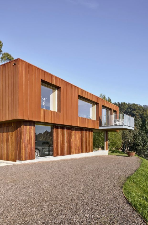 Exterior view of a contemporary two-storey residence with a long, low-pitched roof form and extensive vertical timber cladding. Large recessed windows and glazing punctuate the upper level, while the ground floor includes a partially enclosed garage and timber screening. A projecting upper-level balcony with a metal balustrade overlooks the driveway and landscaped setting.