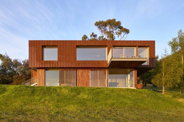 Exterior view of a modern two-storey house set in a landscaped grassy site. The building has a strongly rectilinear form with projecting upper-level volumes, extensive glazing, vertical timber cladding and timber screening, plus a recessed balcony beneath the cantilevered upper floor.